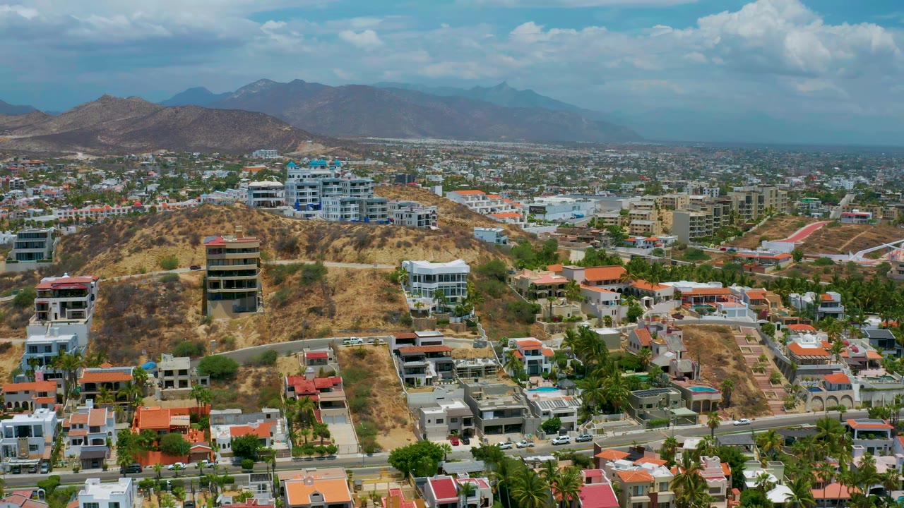 San Jose del Cabo aerial view at 0:01 with the blue-roofed hillside complex near center, residential streets below, mountains on the horizon, and no visible people.