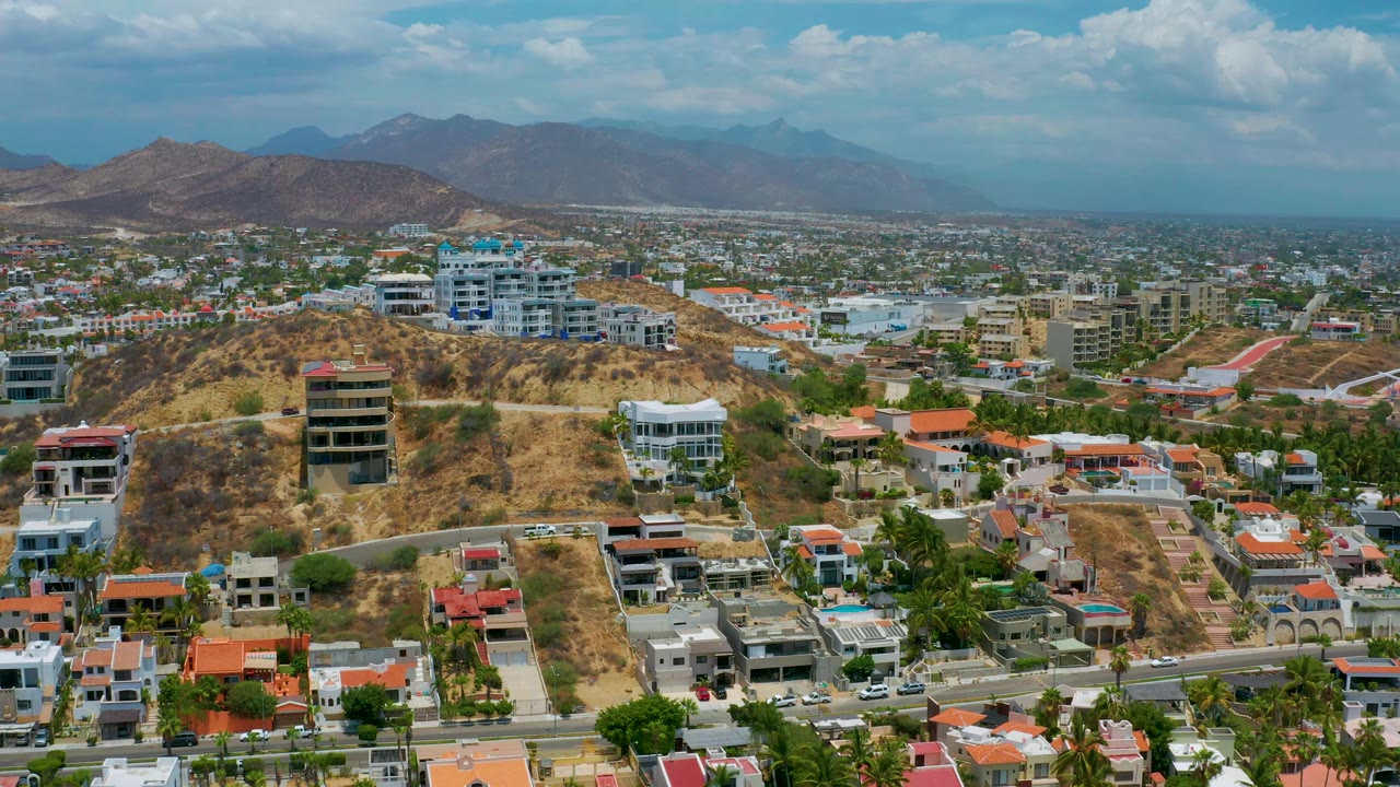 San Jose del Cabo frame at 0:02 shows a wide hillside neighborhood, denser buildings beyond it, hazy mountains in back, scattered vehicles, and no visible people.