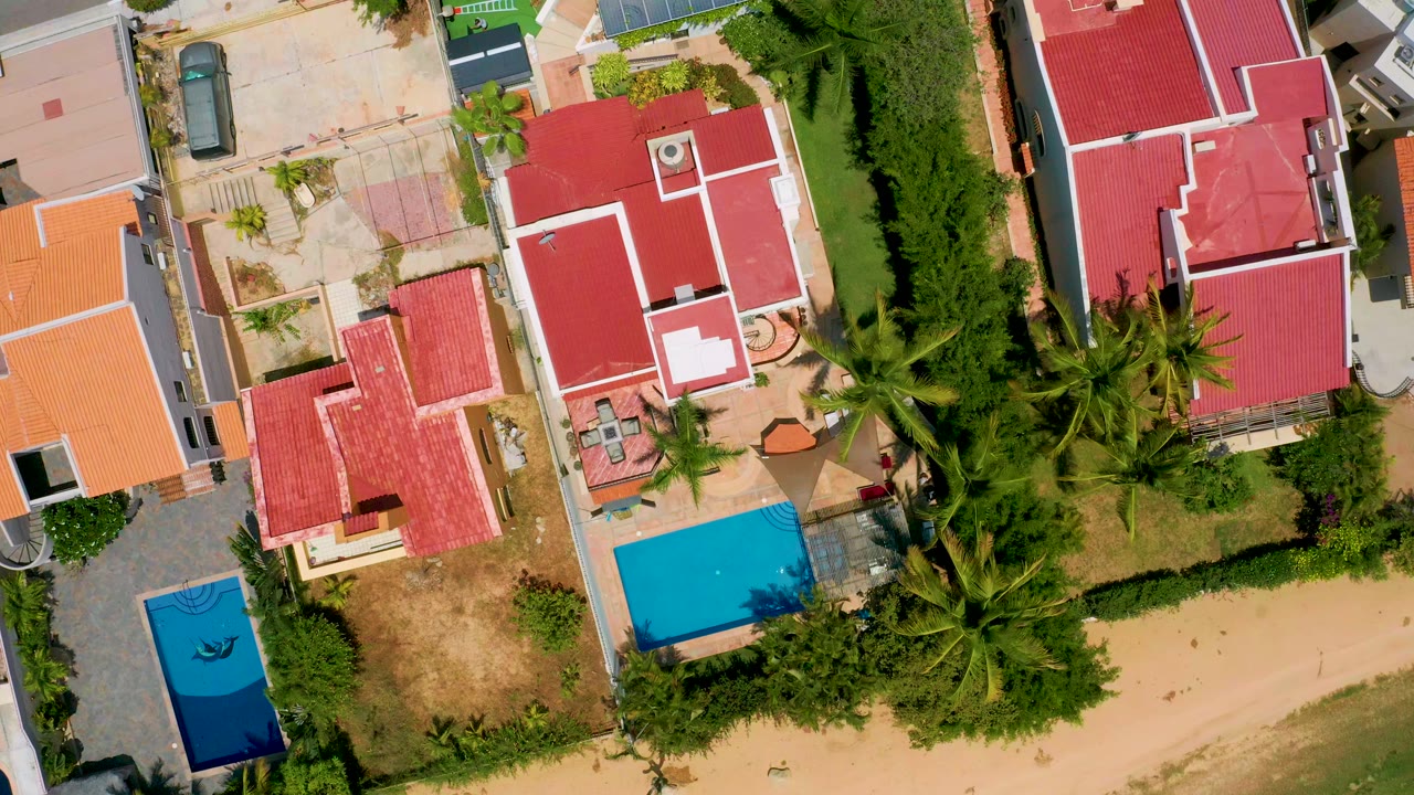 Top-down aerial frame from San Jose del Cabo showing red and orange roofed homes, palms, paved drives, yards, and blue pools with no visible people.