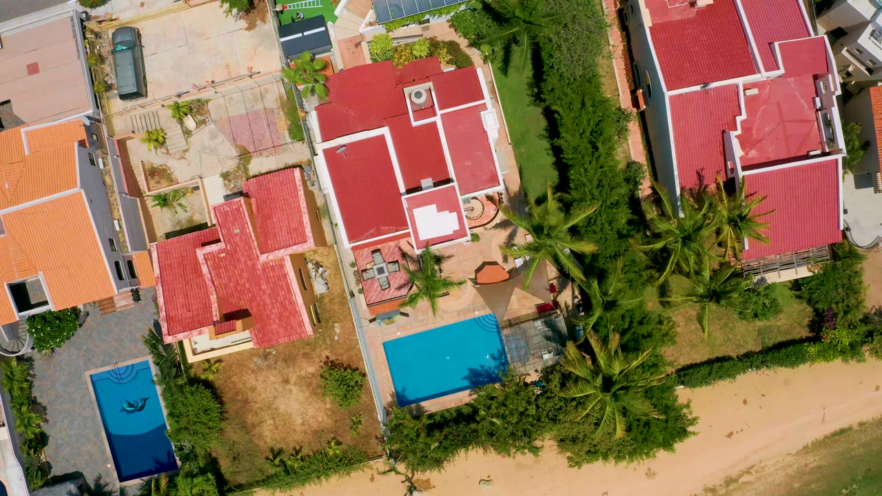 Aerial overhead frame in San Jose del Cabo showing a tighter arrangement of red roofed houses, two blue pools, palms, and paved residential surfaces with no visible people.