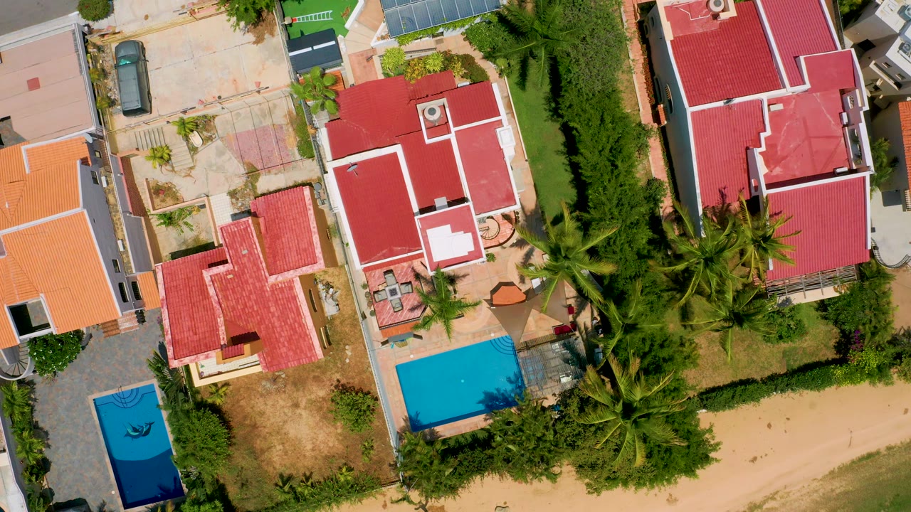 Vertical aerial view over San Jose del Cabo homes with red roofs, bright blue pools, thick palms, small yards, and a sandy road edge, with no visible people.