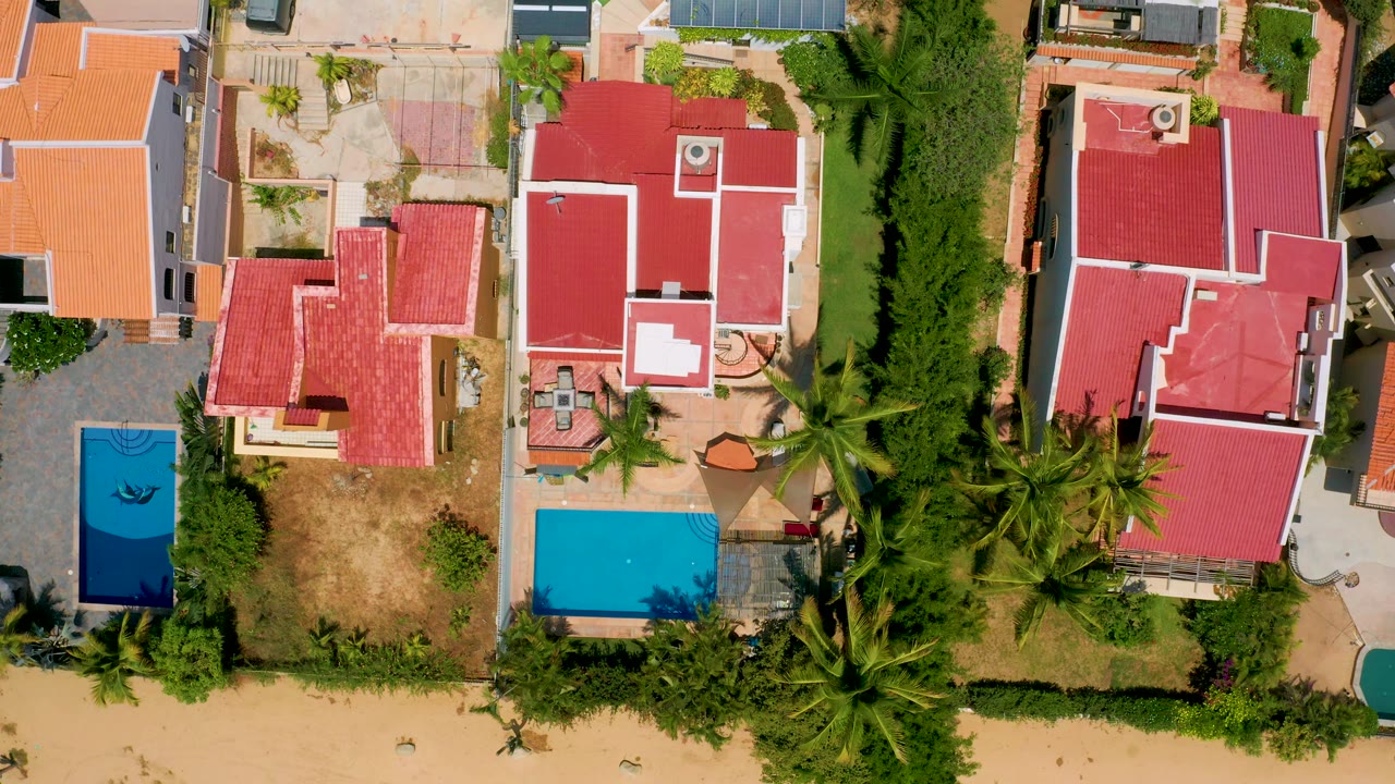 Top-down San Jose Del Cabo residential frame showing red and orange roofs, a turquoise pool near the lower center, a darker blue pool at left, palms, and no visible people.