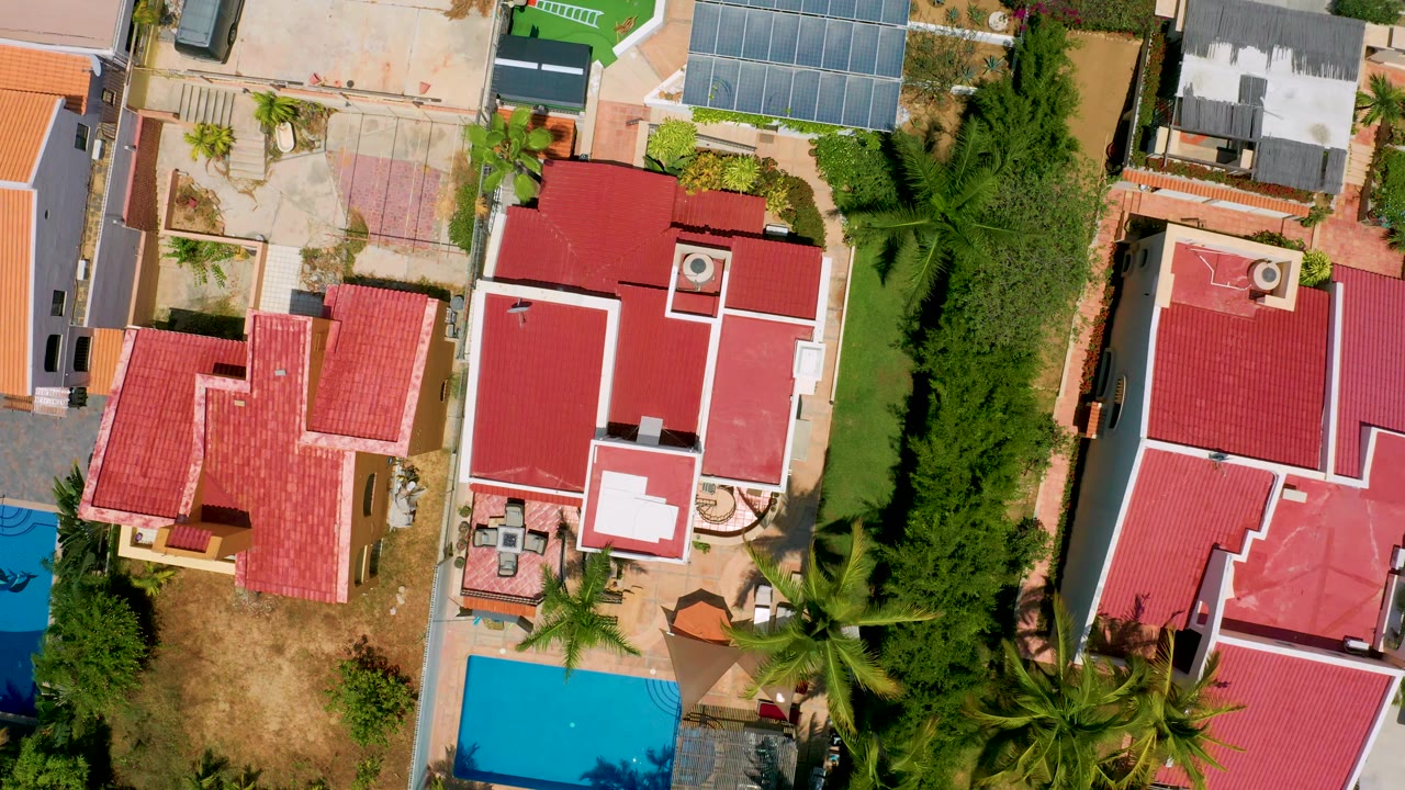 Near-vertical aerial view in San Jose del Cabo with red and terracotta rooftops, a blue pool at frame edge, tall palms, dry grass, and no visible people.