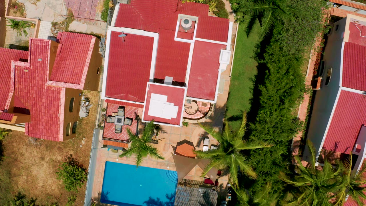 Overhead aerial frame in San Jose Del Cabo showing red roofs with white trim, a narrow green lawn, dense palms at the bottom, and no visible people.