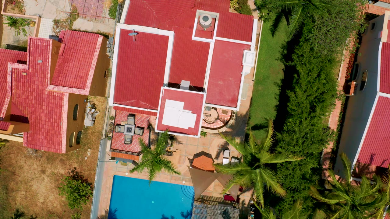 Top-down San Jose Del Cabo frame with red-roof buildings, a partial blue pool at lower left, tall palms along the lower edge, and no visible people.