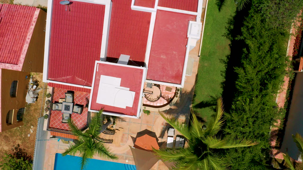 Overhead San Jose Del Cabo frame showing red roof sections, a patio seating area, a pool edge, green lawn, dense hedges, palm fronds, and no visible people.