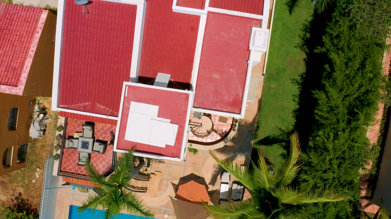 San Jose Del Cabo aerial frame showing red clay-colored roofs, a white rooftop surface, patio chairs, a small pool corner, lawn, tall hedges, and no visible people.