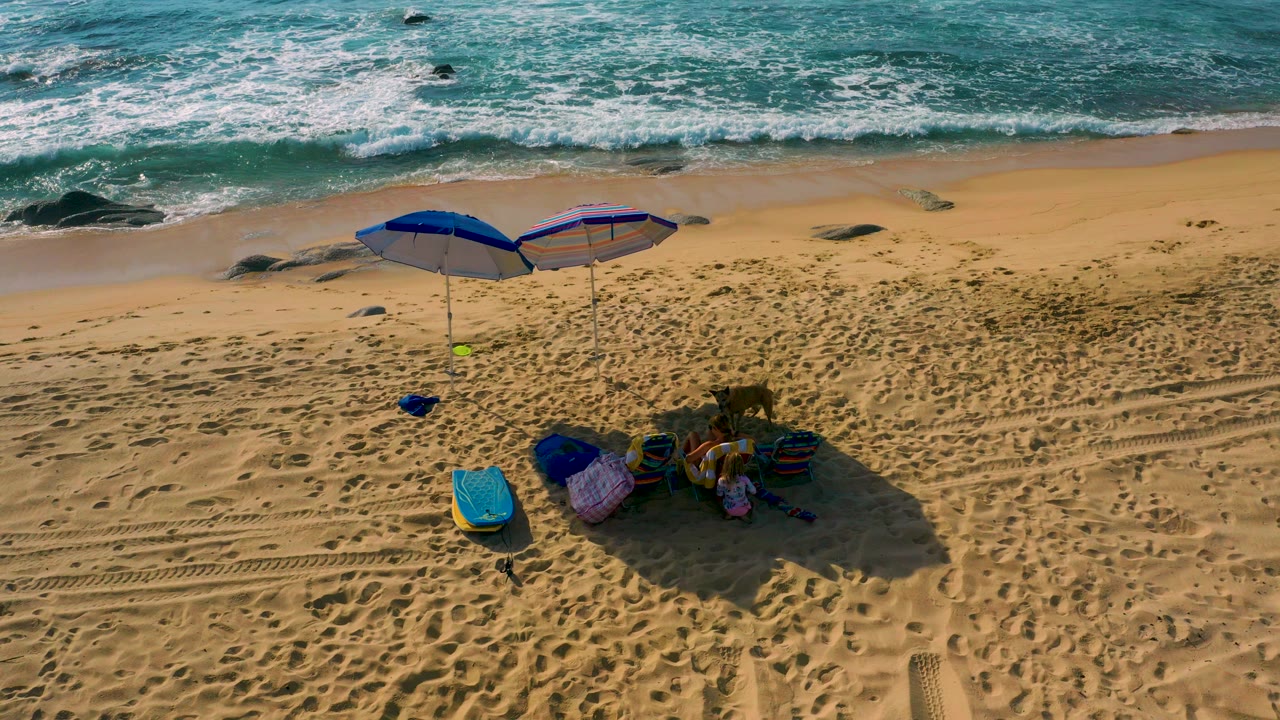 Aerial frame at Shipwrecks Beach in Baja California Sur shows turquoise surf above golden sand, two blue umbrellas, beach gear, long shadows, and no visible people.