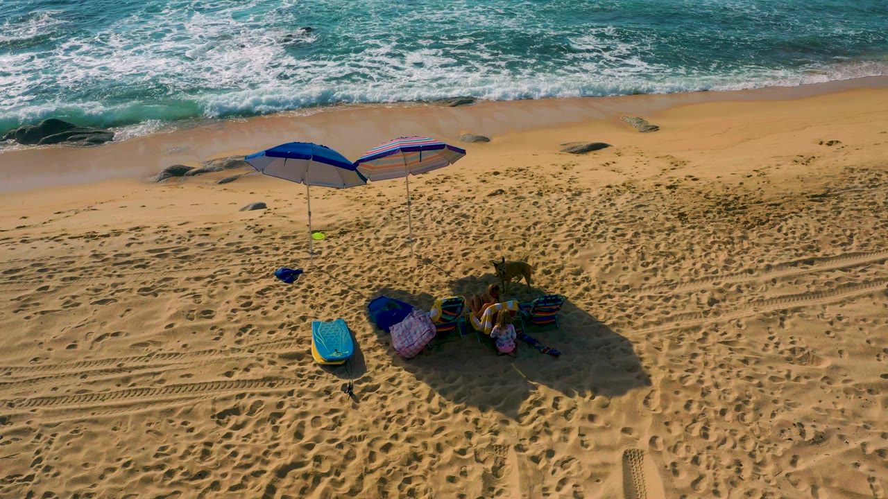 Aerial beach frame in Baja California Sur shows foamy water across the top, two blue umbrellas on open sand, nearby beach items, and no visible people.