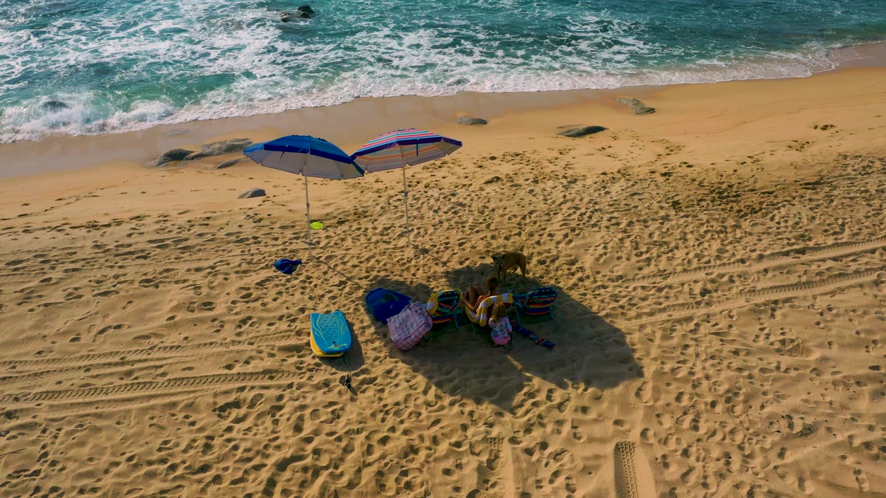 Shipwrecks Beach appears from above with wave foam reaching closer to the sand, rocks near the shore, two blue umbrellas, beach gear, and no visible people.