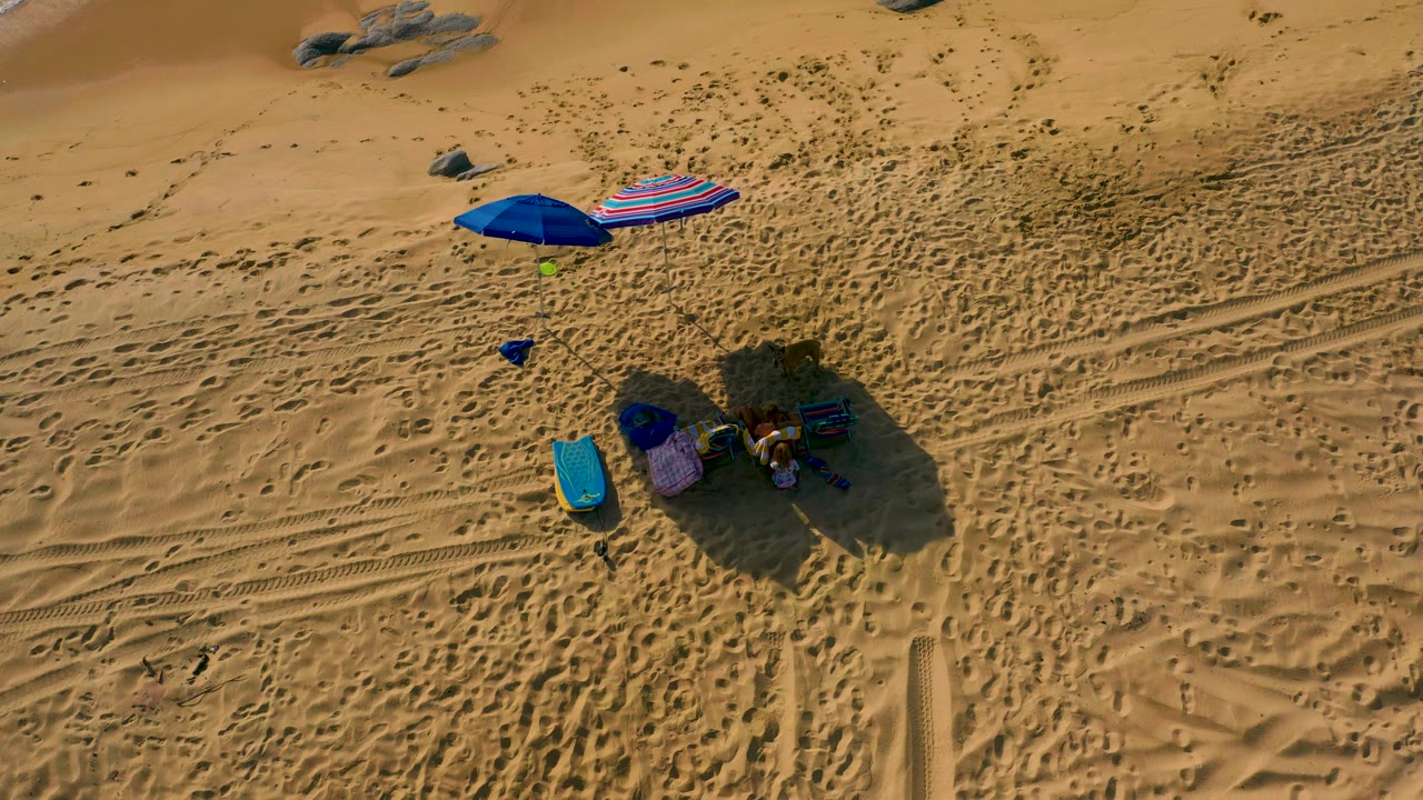High aerial frame from Shipwrecks Beach with the umbrella cluster slightly left of center, long shadows falling across rippled sand, visible footprints and tracks, and small people gathered near chairs and bags.