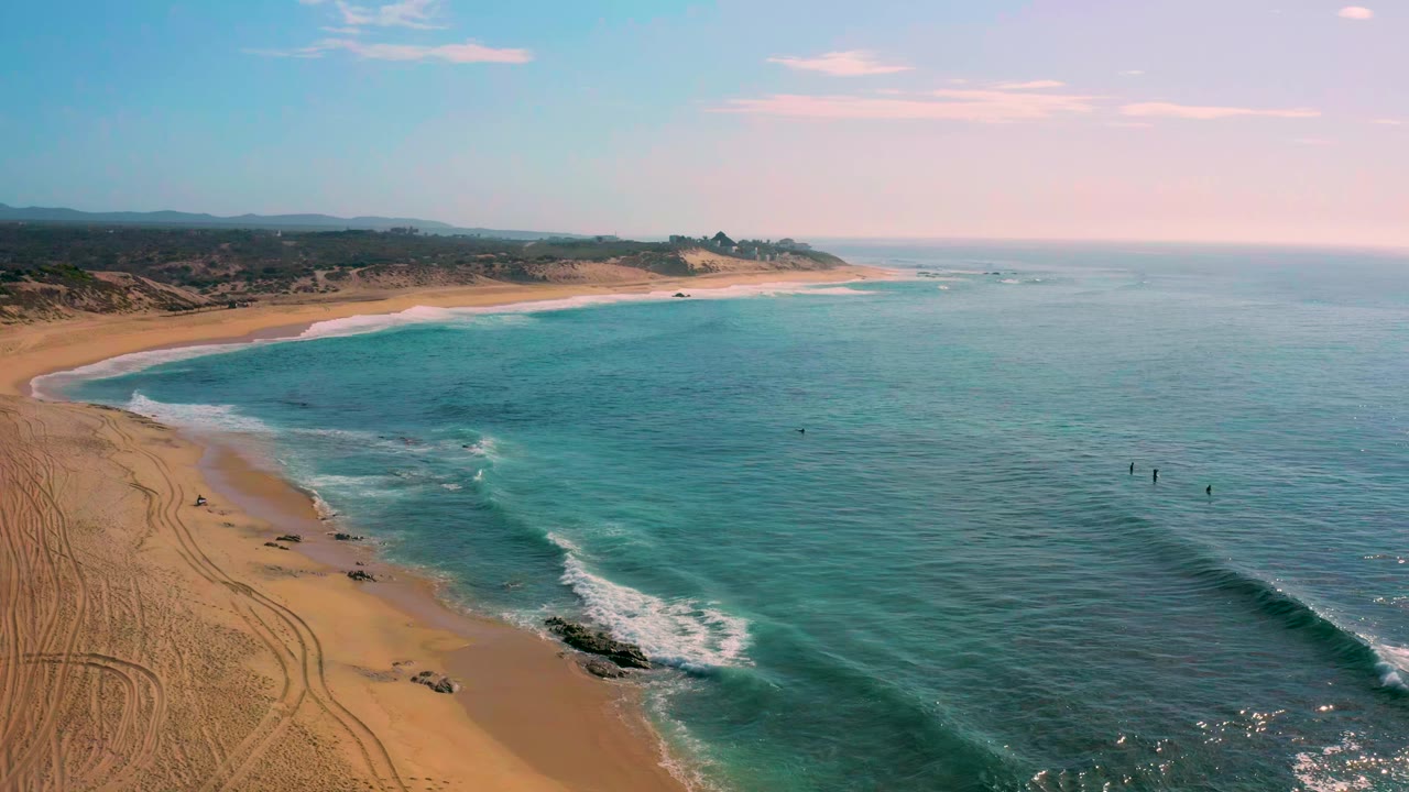 Aerial view at Shipwrecks Beach with the shoreline curving from lower left toward the distance, small white wave breaks near shore, blue-green water filling the right side, and no visible people.