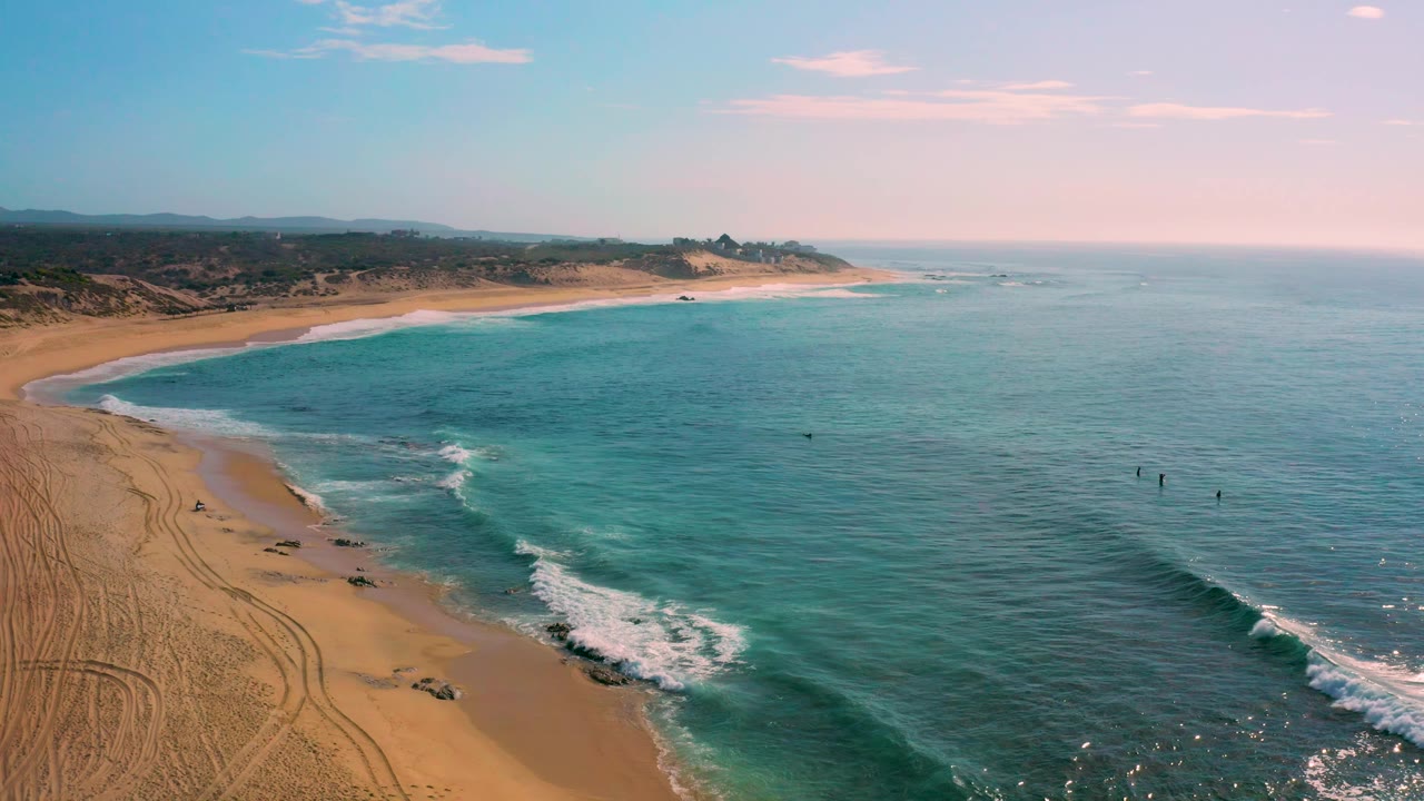 Aerial frame of Shipwrecks Beach showing pale sand at left, clear turquoise shallows through the center, darker open sea at right, small whitecaps offshore, and no visible people.