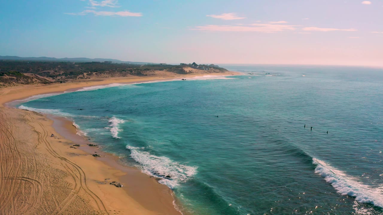 Elevated view over Shipwrecks Beach with a foam band entering the cove from the lower center, open water across the right half, inland dry vegetation beyond the sand, and no visible people.