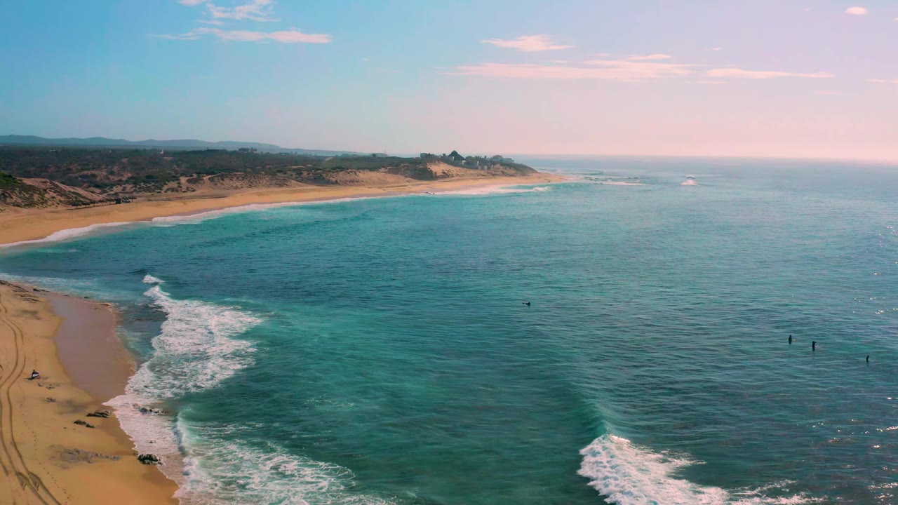Aerial view at Shipwrecks Beach in Baja California Sur, showing a long sand curve, surf foam near the lower left, broad blue-green ocean to the right, and no visible people.