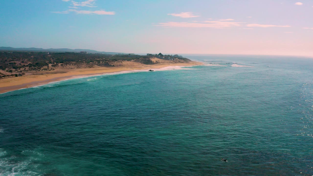 Opening aerial view over Shipwrecks Beach in Baja California Sur, with dark blue foreground water, a golden sand strip on the left, low inland hills, pale clouds, and no visible people.