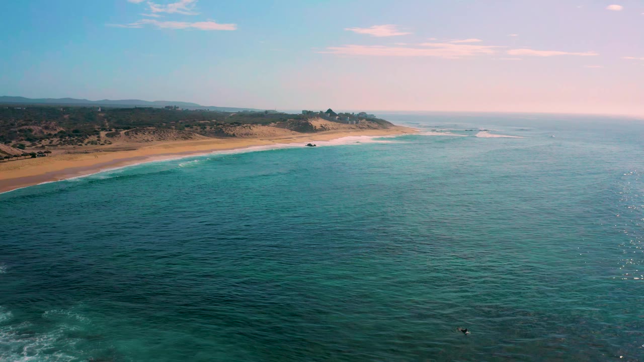 Aerial frame over Shipwrecks Beach showing turquoise shallows near the sand, deeper water across the foreground, low brush inland, a distant shoreline curve, and no visible people.