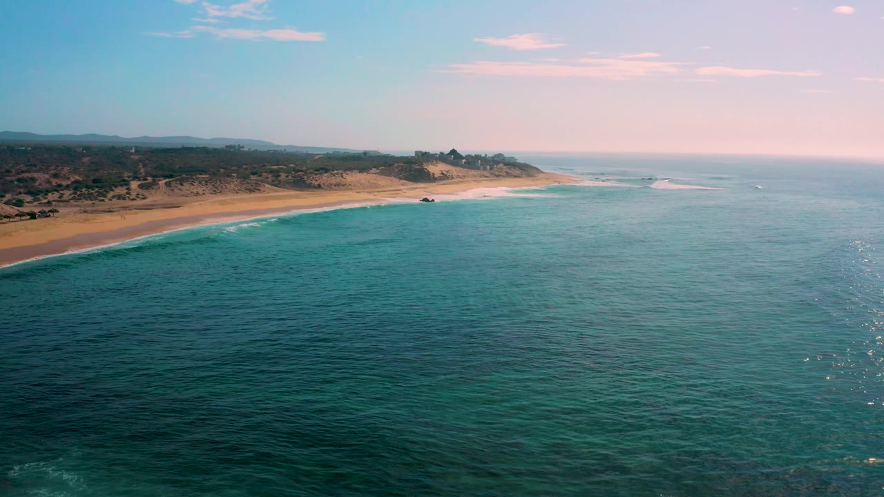 Wide drone view of Shipwrecks Beach with the beach angled across the left side, small white surf breaks offshore, broad blue-green water, hazy inland terrain, and no visible people.