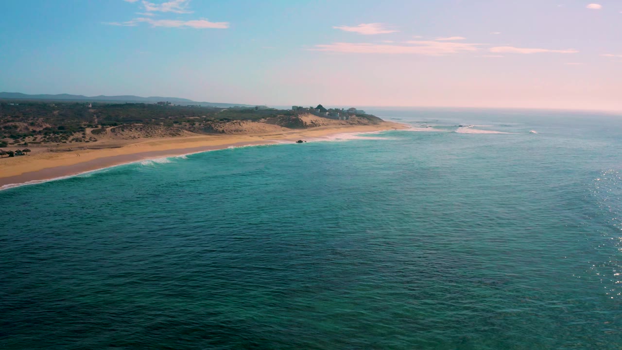 Aerial view of sandy beach and calm blue water with low hills inland.
