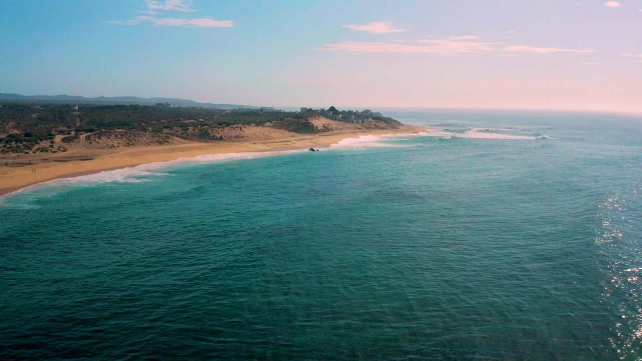 Aerial view of turquoise ocean beside a sandy beach and low coastal hills.