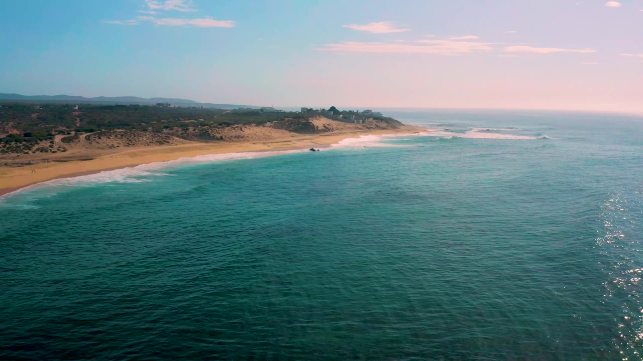 Shipwrecks Beach coastline from above, showing a long strip of sand, gentle white surf, darker hills inland, open ocean to the right, and no visible people.