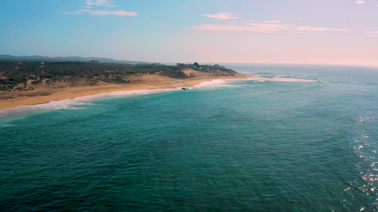Aerial view at Shipwrecks Beach with ocean texture across the foreground, a curved sandy shore on the left, a low cliffed point in the distance, and no visible people.