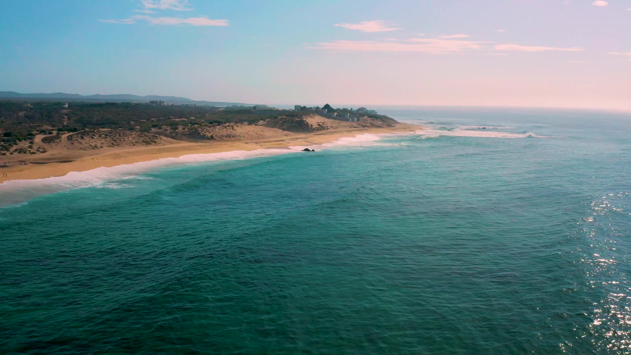 Mid-clip frame over Shipwrecks Beach, with the sandy beach stretching left to right, surf rolling in parallel bands, inland hills in shade, and no visible people.