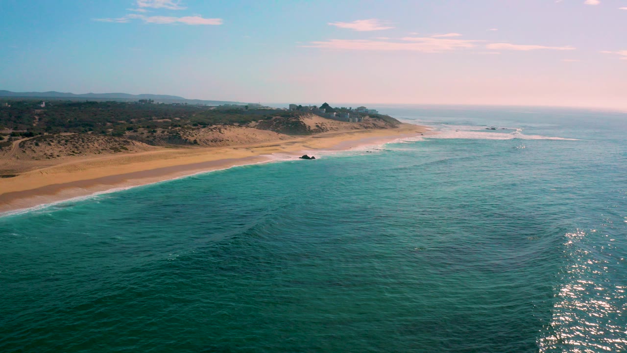 Wide aerial frame over Shipwrecks Beach, Baja California Sur, with turquoise water in the foreground, a long sandy shoreline to the left, low dunes inland, and no visible people.