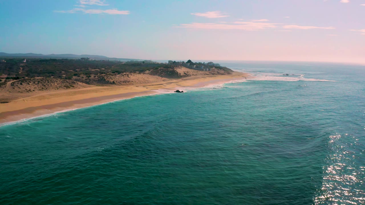 Aerial view at Shipwrecks Beach with sun glinting on the water at the lower right, soft surf along the sand, low dune vegetation inland, and no visible people.
