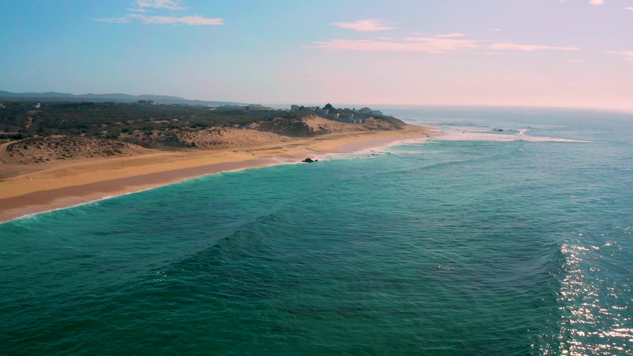High coastal frame at Shipwrecks Beach showing blue-green ocean across most of the image, the beach and dunes on the left, small white breakers offshore, and no visible people.