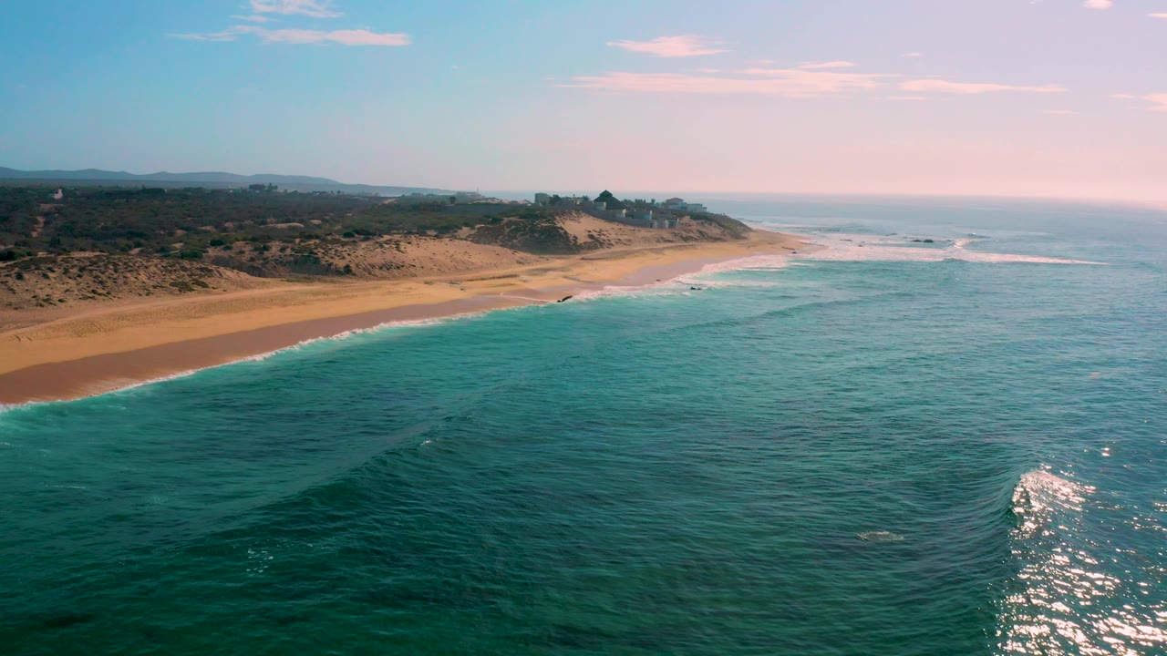 Shipwrecks Beach appears from above with a clean curve of white foam along the sand, darker green water in the foreground, dunes behind the beach, and no visible people.