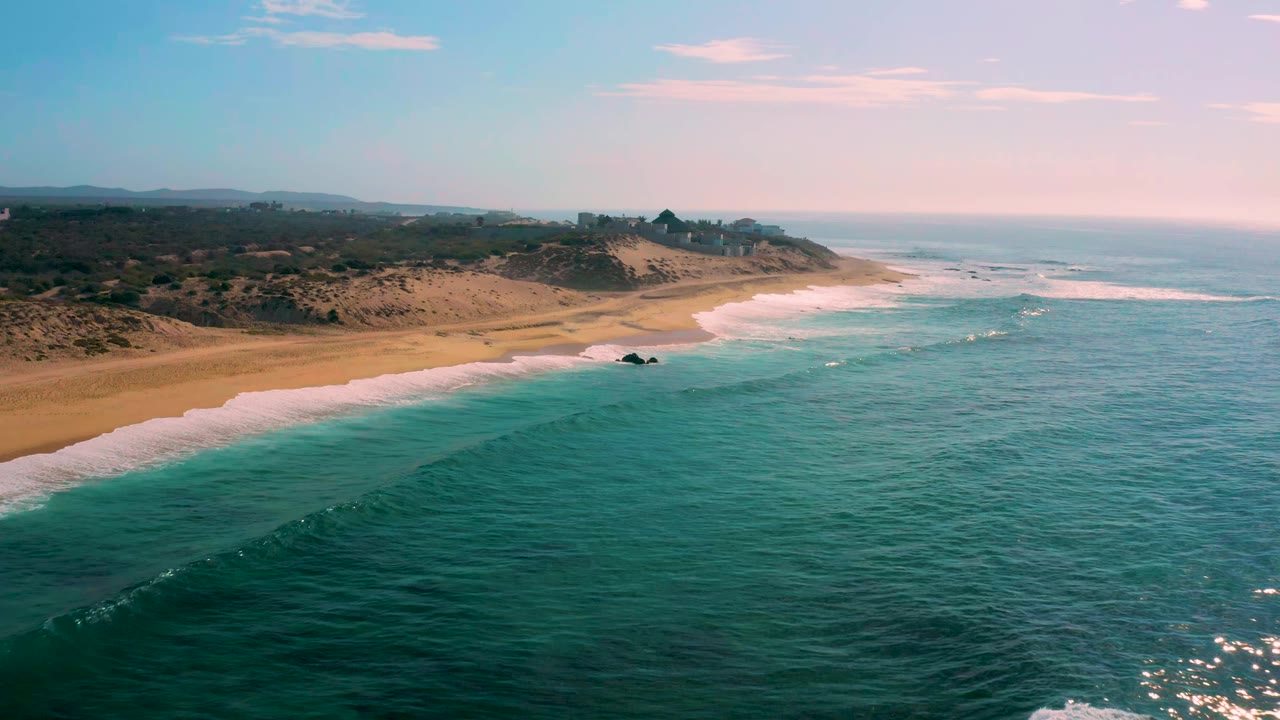 Aerial frame at Shipwrecks Beach, Baja California Sur, showing white surf curling toward the sand, darker scrubland behind the dunes, and no visible people.