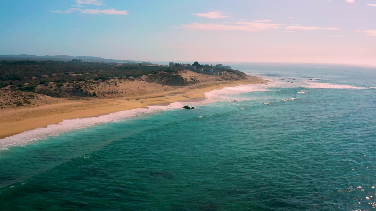 Aerial view along Shipwrecks Beach in Baja California Sur, with more open water filling the lower frame, surf breaking near shore, dunes inland, and no visible people.