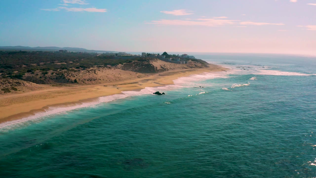 Mid-clip aerial frame over Shipwrecks Beach, Baja California Sur, where a small dark rock sits near the surf line, with broad sand and scrub dunes behind it and no visible people.