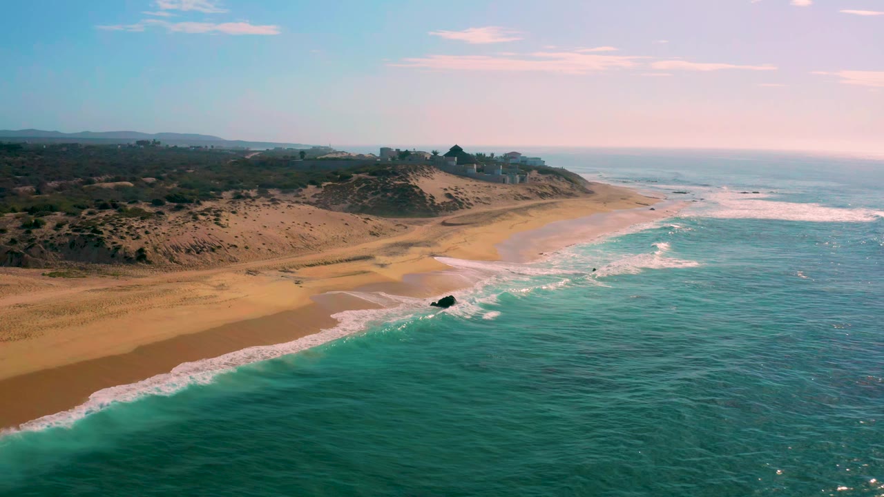 Wide aerial frame of Shipwrecks Beach, Baja California Sur, showing white waves brushing the shoreline, open tan sand, scrub-covered dunes, small distant buildings on the bluff, and no visible people.