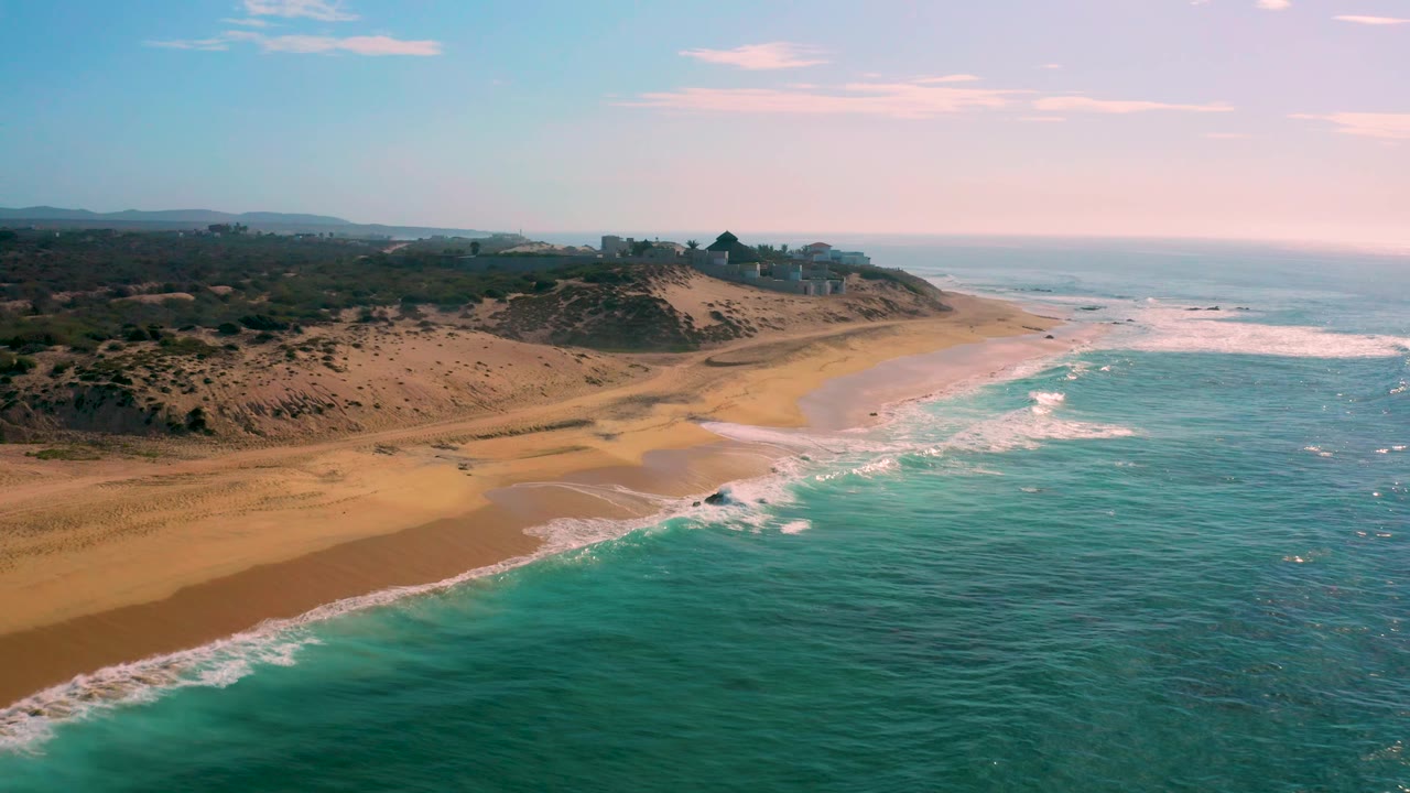Aerial shoreline view at Shipwrecks Beach in Baja California Sur, with teal ocean in the foreground, white foam reaching the beach, dune ridges to the left, distant coastal structures, and no visible people.