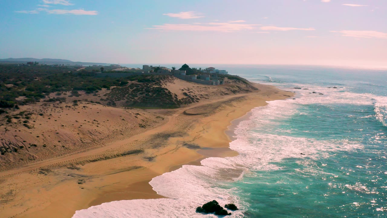 Aerial view at Shipwrecks Beach in Baja California Sur, Mexico, with turquoise surf entering from the right, tan beach below, scrubby dunes at left, distant ridge structures, and no visible people.