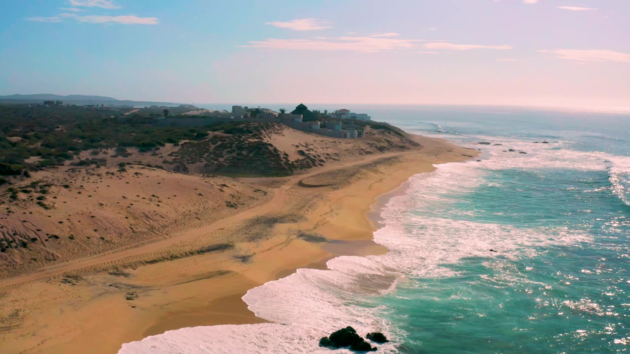 Aerial frame at Shipwrecks Beach showing foamy waves pushing farther up the right edge of the beach, a broad tan shoreline, darker dune scrub to the left, ridge buildings in the distance, and no visible people.