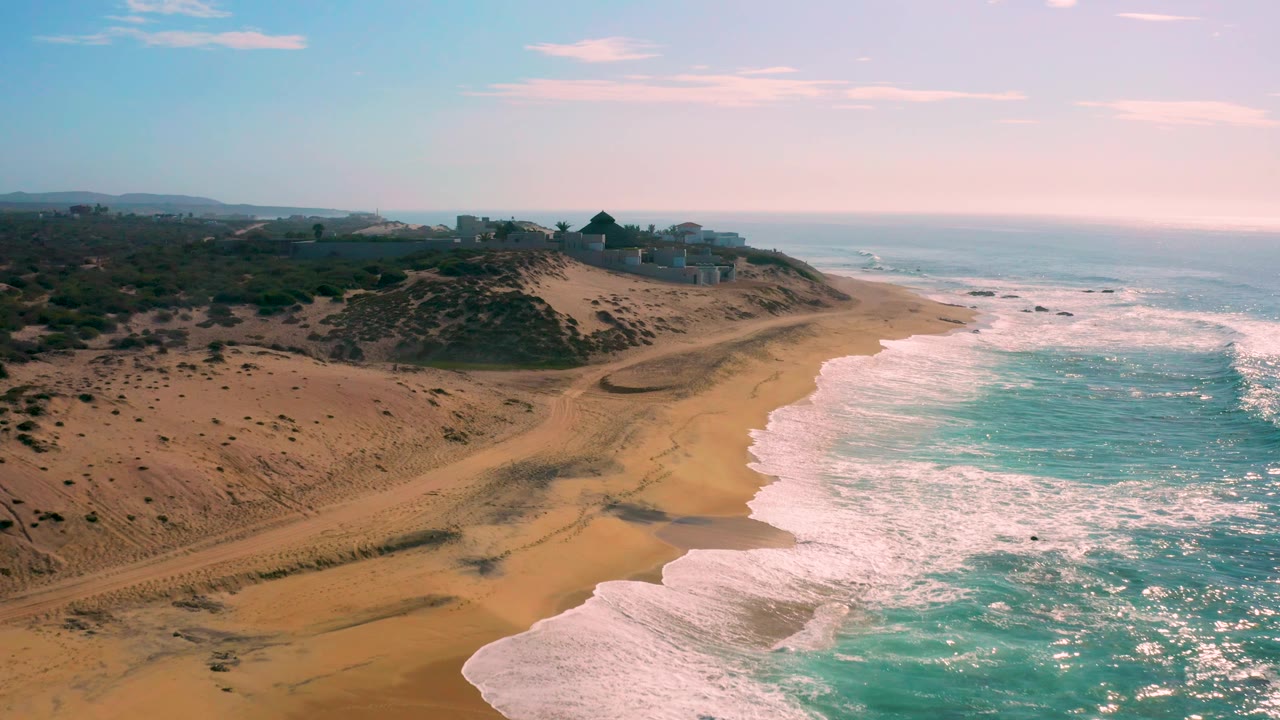 Aerial view over Shipwrecks Beach with sparkling turquoise water on the right, white surf bands crossing the sand, vehicle-like tracks or paths in the dunes, distant ridge structures, and no visible people.