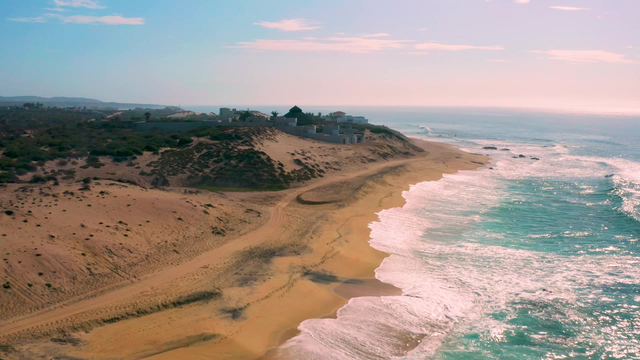 Aerial frame at Shipwrecks Beach where the shoreline curves through the center, foamy water fills the lower right, scrub-covered dunes fill the left, distant coastal buildings sit on the ridge, and no visible people.