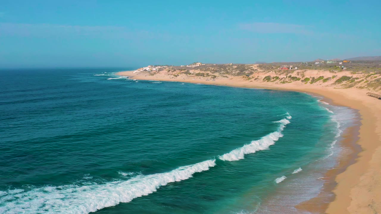 Wide aerial frame of Shipwrecks Beach showing deep blue water at left, a pale turquoise shallows band, white foam nearing the sand, dune vegetation, and no visible people.