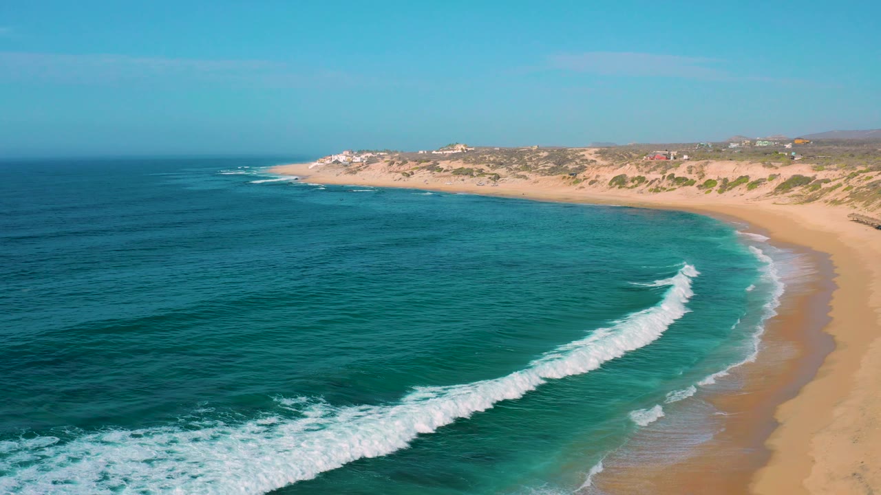 Aerial view of Shipwrecks Beach with a longer white breaker arcing through green-blue water toward the right-side sand, backed by low dunes and no visible people.