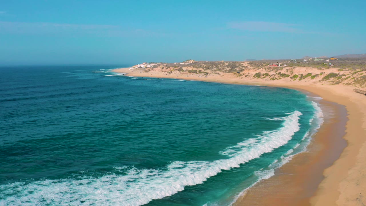 Shipwrecks Beach seen from above, with darker ocean in the lower left, turquoise water near shore, a thin white wave line, tan beach, dunes, and no visible people.
