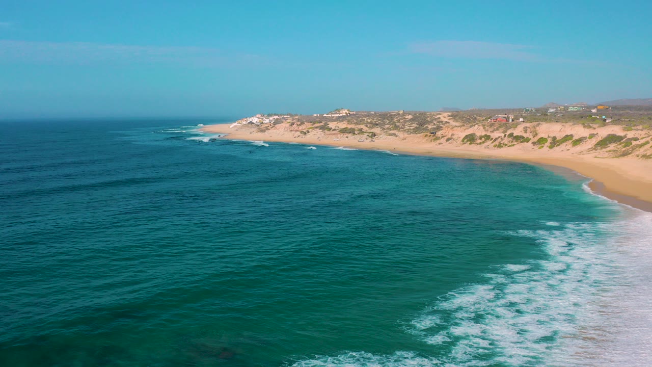 Opening aerial frame over Shipwrecks Beach, Baja California Sur, with teal water in the foreground, a pale sand beach and scrub-covered dunes beyond, and no visible people.