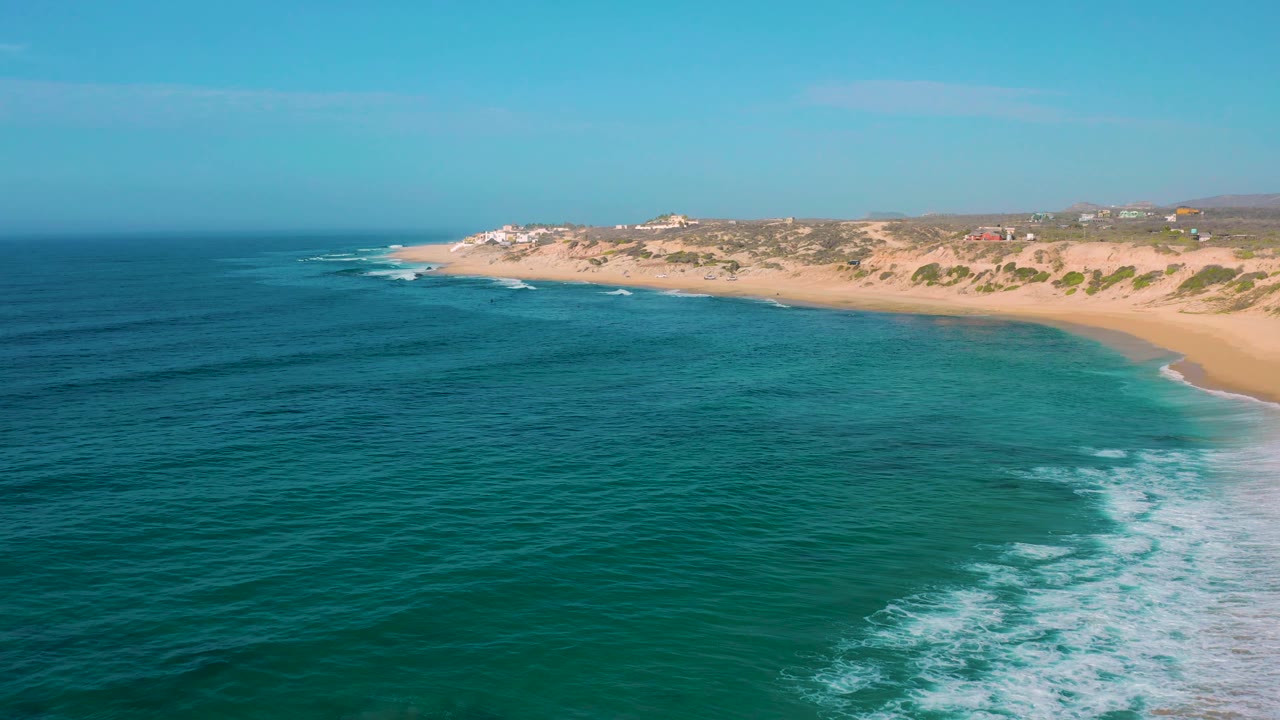 Shipwrecks Beach aerial frame with darker ocean bands in the foreground, pale turquoise shallows near the shore, a sweeping sand beach, scrubby dunes, and no visible people.