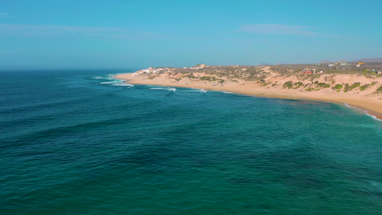 Wide aerial frame of Shipwrecks Beach in Baja California Sur, with blue-green water in the foreground, tan sand along the right side, low scrub behind the beach, and no visible people.