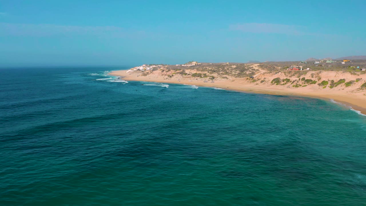 Aerial view over Shipwrecks Beach with darker ocean bands in the foreground, pale surf lines reaching the sand, scrub-covered dunes beyond, and no visible people.