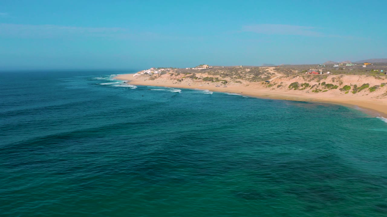 Shipwrecks Beach appears from above with turquoise water spreading across the foreground, a thin strip of white surf at the shore, sandy dunes in back, and no visible people.