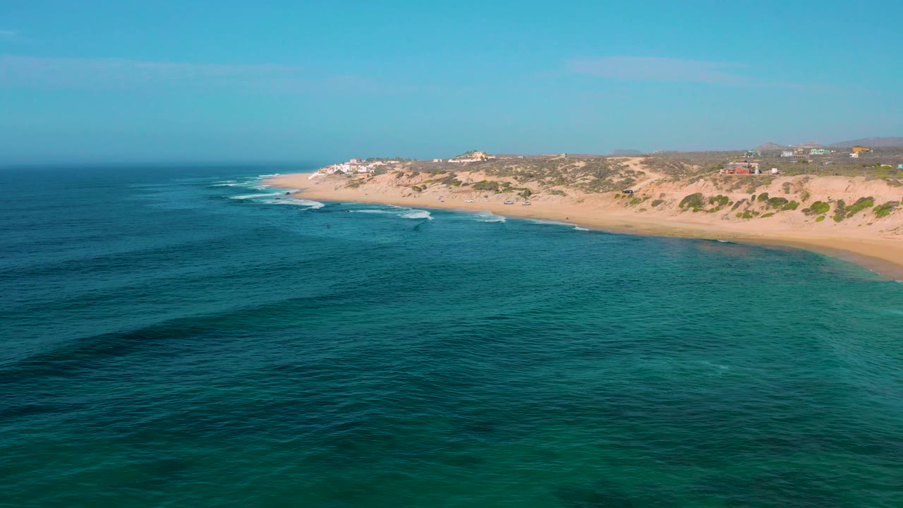 Aerial frame over Shipwrecks Beach with broad ocean foreground, small waves rolling toward the empty sand, low vegetation behind the beach, and no visible people.
