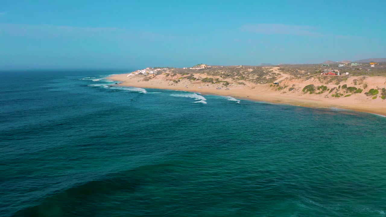 Shipwrecks Beach shoreline seen from above with white wave streaks approaching the sand, low vegetation behind the beach, distant structures, and no visible people.